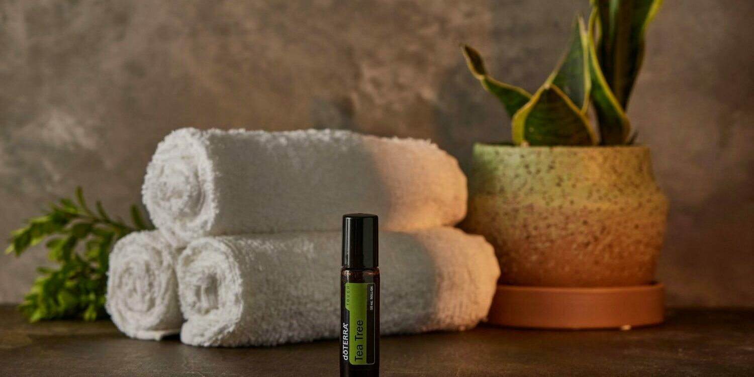 A green essential oil bottle sits in front of rolled white towels and a green plant.